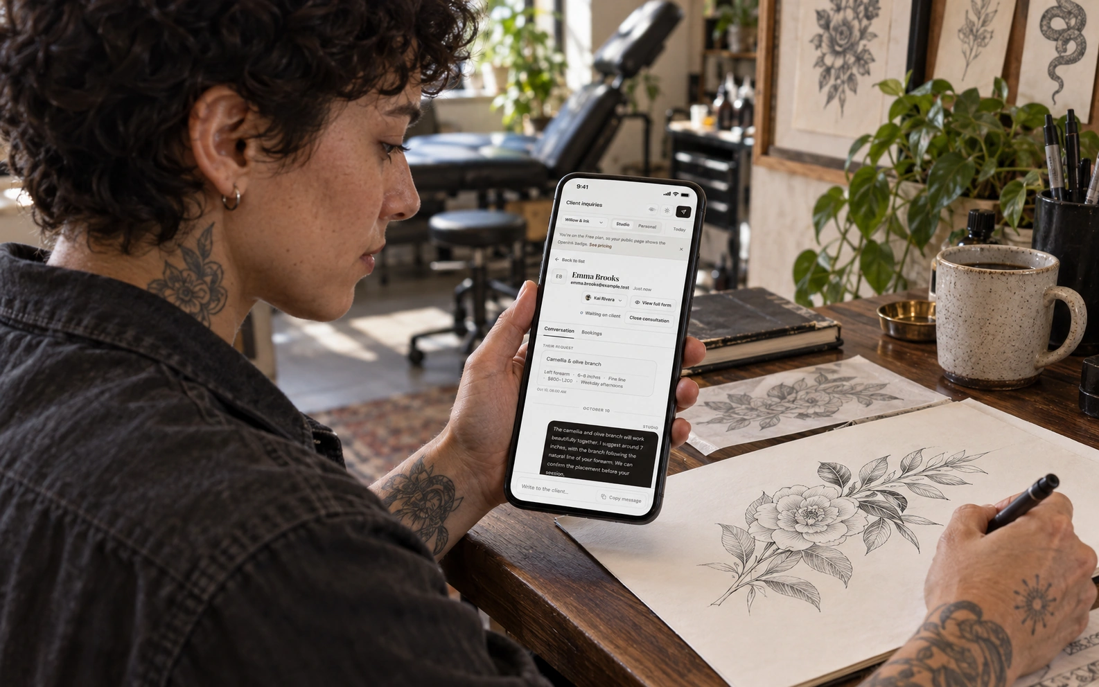 Tattoo artist reading an English client inquiry on a phone while preparing a botanical sketch